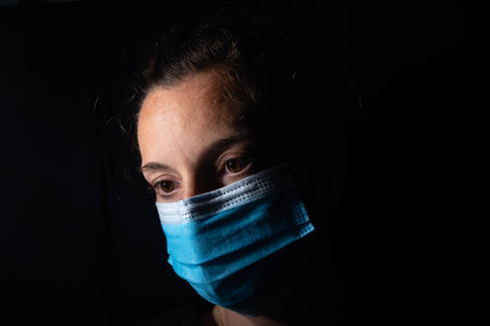 Close-up of a dark portrait of a very dimly lit woman. The woman is wearing a surgical mask, her gaze is lost on the horizon and she transmits feelings of sadness or hopelessness. Depression during the coronavirus.の写真素材