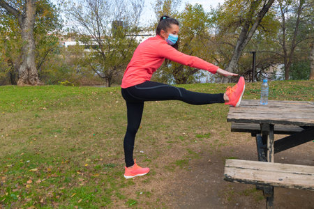 Sporty dressed woman stretches her legs on a picnic table in a park wearing a surgical mask to...