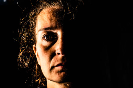 Dark and very sharp portrait of a serious woman with only half her face lit up on a black background looking sad or in a depression. Very hard look on her face.の写真素材