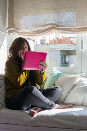 Caucasian woman aged 50 looking at a tablet with concern. Sitting on a informal way on a sofa close to the window. COVIDの写真素材