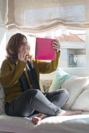 Caucasian woman aged 50 during a video chat at home. Brown hair, sitting on a sofa in an informal way, greeting the audience.の写真素材