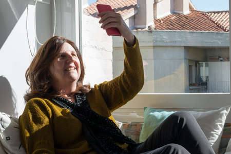 Caucasian woman aged 50 using the phone at home for a video chat during COVID. Sitting on a sofa near the window, sun comming through the glass.の写真素材