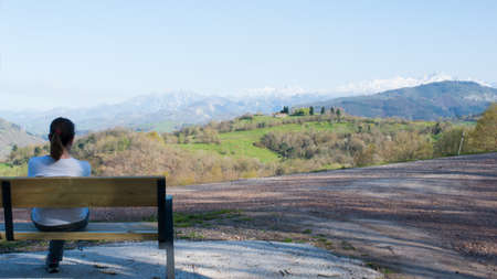 Young woman seen from her back sitting alone on a wooden bench, looking to a beautiful green landscape on a sunny day. Picos de Europa mountains with snow in the background.の写真素材