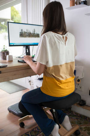 Young woman working at home using an ergonomic kneeling chair seen from her back.の写真素材