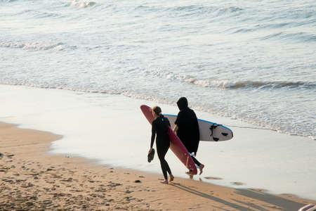 Young couple walking on the beach with their surf boards looking for the right spot to start surfing. Sunset, San Lorenzo beach, Gijon, Asturias, Spain. Europeの写真素材