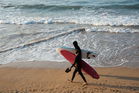 Young couple walking on the beach with their surf boards and wetsuits. Sunset, San Lorenzo beach,...