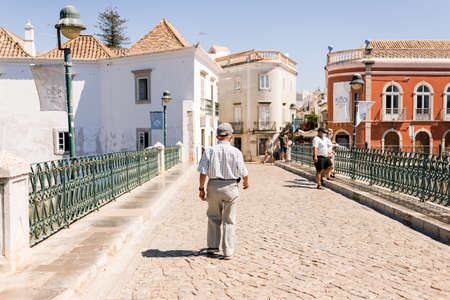 Tavira, Portugal; August 10, 2020: Older man with hygienic mask walking on the touristy Roman bridge of Tavira in the Portuguese Algarve.のeditorial素材