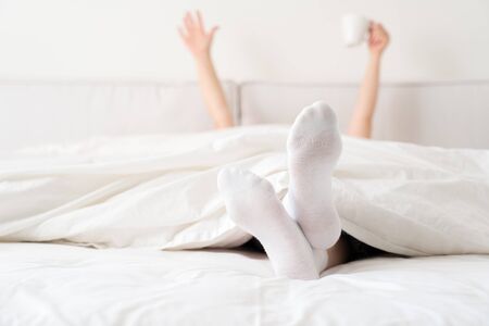 Female hand holding cup of coffee from under a blanket in bed. Woman waking up in the morningの写真素材