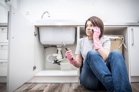 Scared Woman sitting near leaking sink in laundry room calling for helpの写真素材