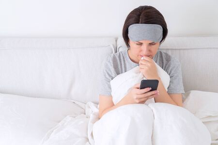 Woman wearing sleeping mask texting on smartphone as she sits in bed covered with duvetの写真素材