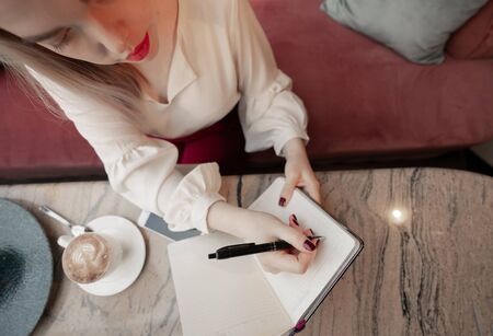 Dreamy young woman writing down future plans and goals in own diary resting in cozy cafeteriaの写真素材