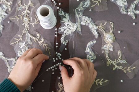 closeup of female hands of seamstress tailor sewing wedding dress using beads and lace in the studio