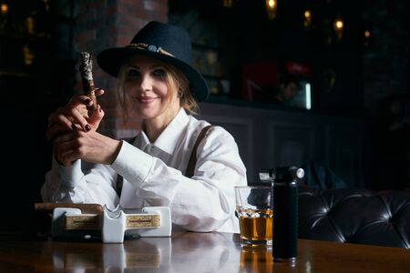 Senior attractive female gangster smoking cigar over dark background in restaurantの写真素材