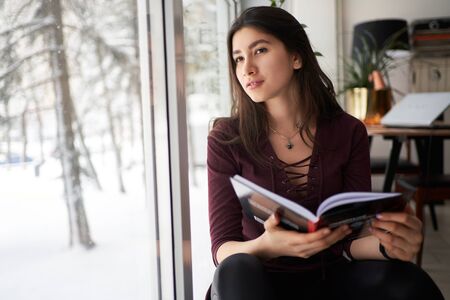 Happy young woman reading a book by a window in winterの写真素材