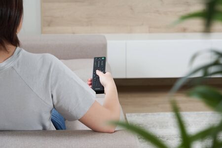 Rear view of young woman watching television and holding remote control while sitting on a couch
