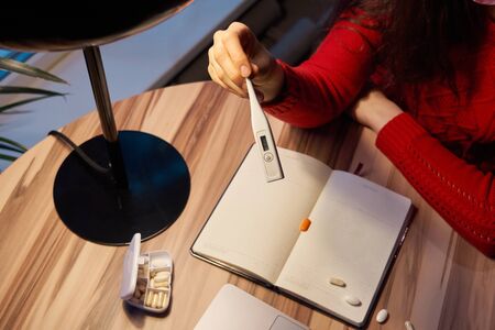 sick woman with pills and thermometer on a table working on a laptop from home on quarantine isolation.の写真素材
