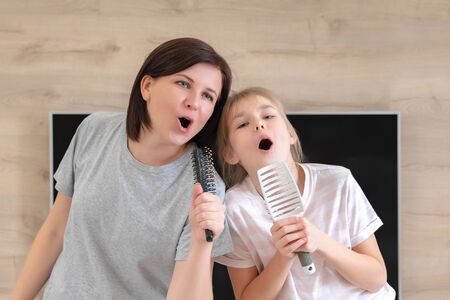 Happy family young adult mother and cute teen daughter having fun singing karaoke song in hairbrushes. mother laughing enjoying funny lifestyle activity with teenage girl at home together.の写真素材