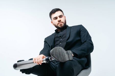 Disabled young man with prosthetic leg sitting on a chair in studio over white background,...