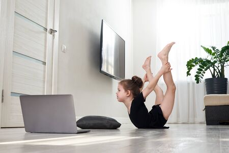 Little girl doing gymnastics exercises at home using online learning with laptop computer, internet education conceptの写真素材