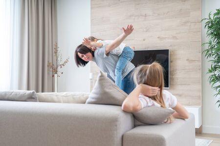 Preteen sad girl sitting on couch while mother having fun with younger sister, jealousy conceptの写真素材