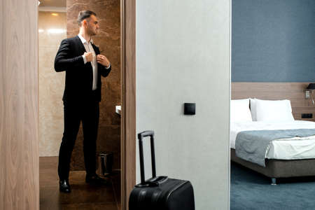 Young traveler businessman wearing white shirt standing at the mirror and getting dressed for meeting or work after arriving in the hotel room with his luggageの写真素材