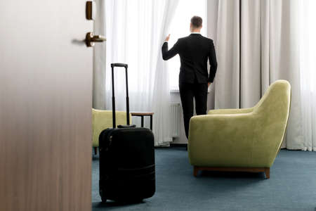 Rear view of businessman wearing dark suit standing in hotel room looking into windowの写真素材