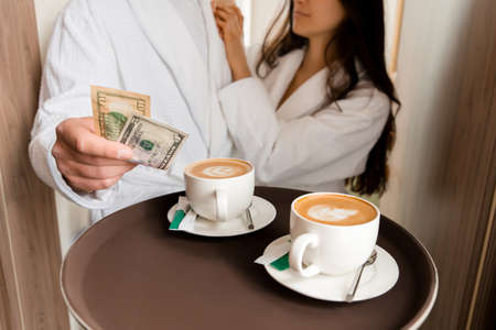 Room service delivering coffee to a hotel room for married couple wearing bathrobe who is giving dollar tipsの写真素材
