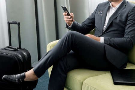 Cheerful businessman using phone sitting in hotel room with suitcase on business trip