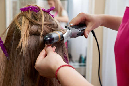 Hairdresser makes hairstyle girl with long hair in a beauty salon. Create curls with curling ironsの写真素材