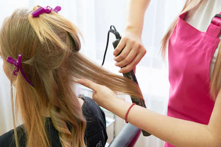 Hairdresser makes hairstyle girl with long hair in a beauty salon. Create curls with curling ironsの写真素材