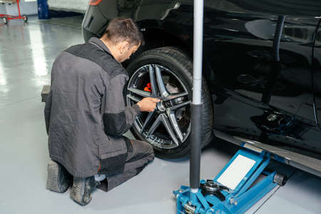 Mechanic changing car wheel in auto repair shop using lift jack and electric drill to loosen the...