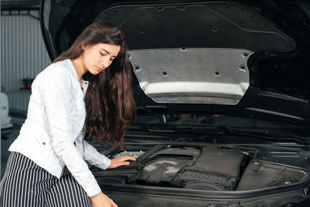 Young woman standing in front of the car with opened hood in garage looking under car hoodの写真素材