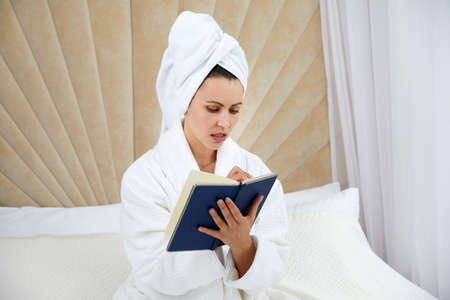 Young woman sits on bed wearing bathrobe writing notes in diary. Blogger creats publication for blog in notebookの写真素材