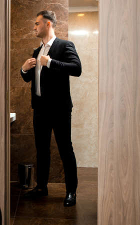 Young traveler businessman wearing white shirt standing at the mirror and getting dressed for meeting or work after arriving in the hotel room with his luggageの写真素材