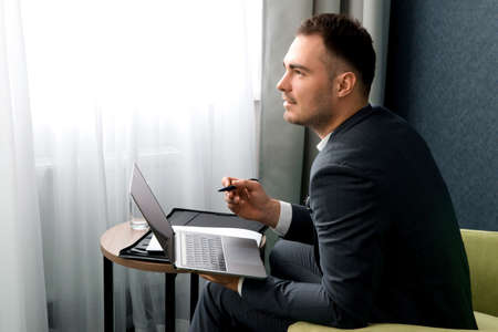 Young businessman is using laptop while sitting at the hotel room with suitcase.の写真素材