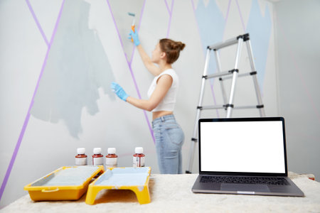 Young woman painting wall with paint roller and using masking tape. Laptop standing on table in front with white copy space mockupの写真素材