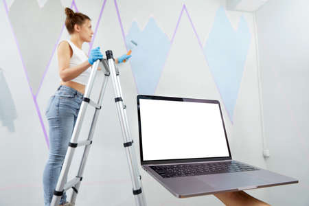 Young woman painting wall with paint roller and using masking tape standing pn ladder. Hand holding laptop in front with white copy space mockupの写真素材