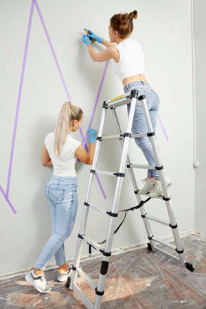 two Young woman preparing wall for painting standing on ladder applying masking tape on wall. Diy projectの写真素材