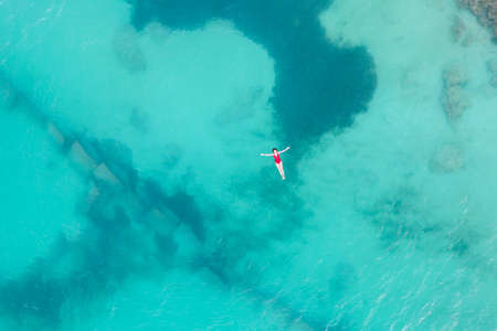 Aerial top view of woman wearing red swimsuit lying on transparent turquoise water surface on caribbean beach. Travel and vacations concept. Tropical background with copy spaceの写真素材