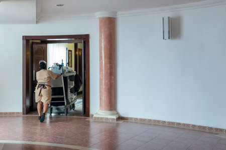 Chambermaid pushing cart along corridor in hotel. Maid at work and trolley with room supplies and cleaning equipment in hotelの写真素材