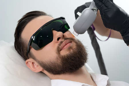Young bearded man getting laser facial treatment by professional cosmetologist in a beauty clinic. Healthy man lifestyle conceptの写真素材