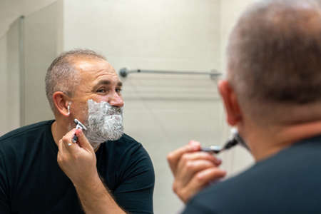 Middle aged handsom man trimming his beard and mustache standing in bathroom and looking in a mirrorの写真素材