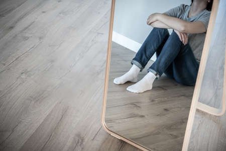 Reflection in mirrow of a sad woman sitting on a floor alone in empty room, despair and lonely concept with copy space, grey toneの写真素材