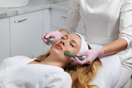 Young woman having facial massage with stone sticks in spa salon