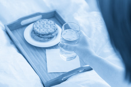Female hand holding glass of water over tray with crackers on bedの写真素材
