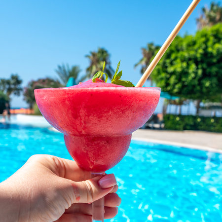 Hand with chilled raspberry Daiquiri and mint leaves by pool on sunny day. Concept of summer relaxation and enjoymentの写真素材