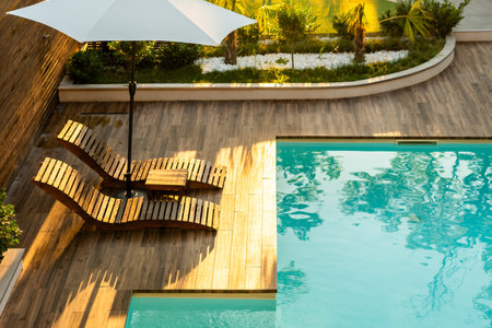 Elegant poolside wooden deck with curved lounge chairs, umbrella, and turquoise swimming pool in...