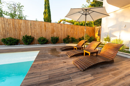 Modern wooden poolside deck with curved lounge chairs, umbrella, and turquoise pool in a sunny backyardの写真素材