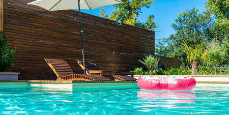 Modern backyard pool with turquoise water, pink inflatable ring, wooden deck, umbrella, and lounge chairsの写真素材