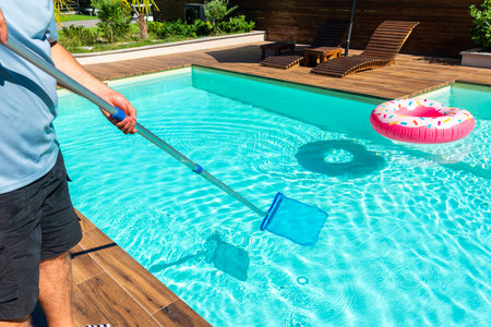 Man cleaning turquoise swimming pool with skimmer net, wooden deck, and colorful inflatable ringsの写真素材